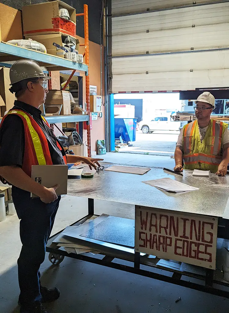 Two men speaking in warehouse setting with table and shelving in the background and an exit to the outdoors