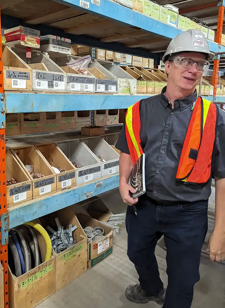 Man in hardhat and orange vest standing in warehouse with various items on shelves in the background