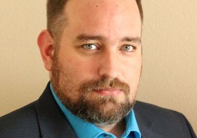 Professional headshot of a man with a trimmed beard wearing a navy blazer and teal shirt, facing forward.